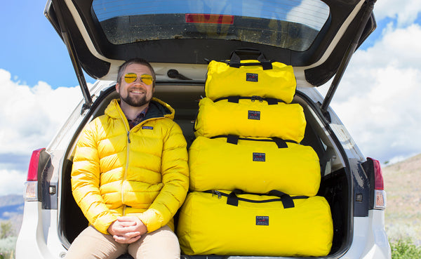 Person in a yellow down jacket sitting in the back of a car with the hatch open. Next to them are stacked Yeomans of each size in Canary.